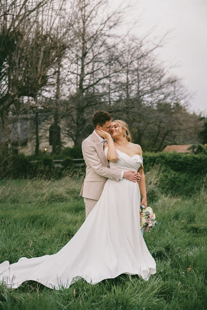 Film wedding photograph capturing an intimate embrace between a newlywed couple in a field at Cossars Wineshed in Christchurch, highlighting timeless tones and organic storytelling.