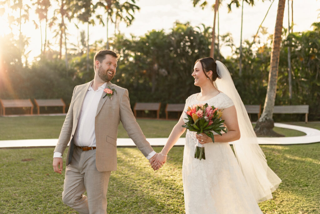 Bride and groom holding hands, smiling at each other, walking in a sunny tropical garden with palm trees.