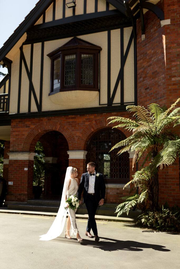 A stylish bride and groom walking hand-in-hand in front of a historic, brick and timber-framed building in Christchurch.