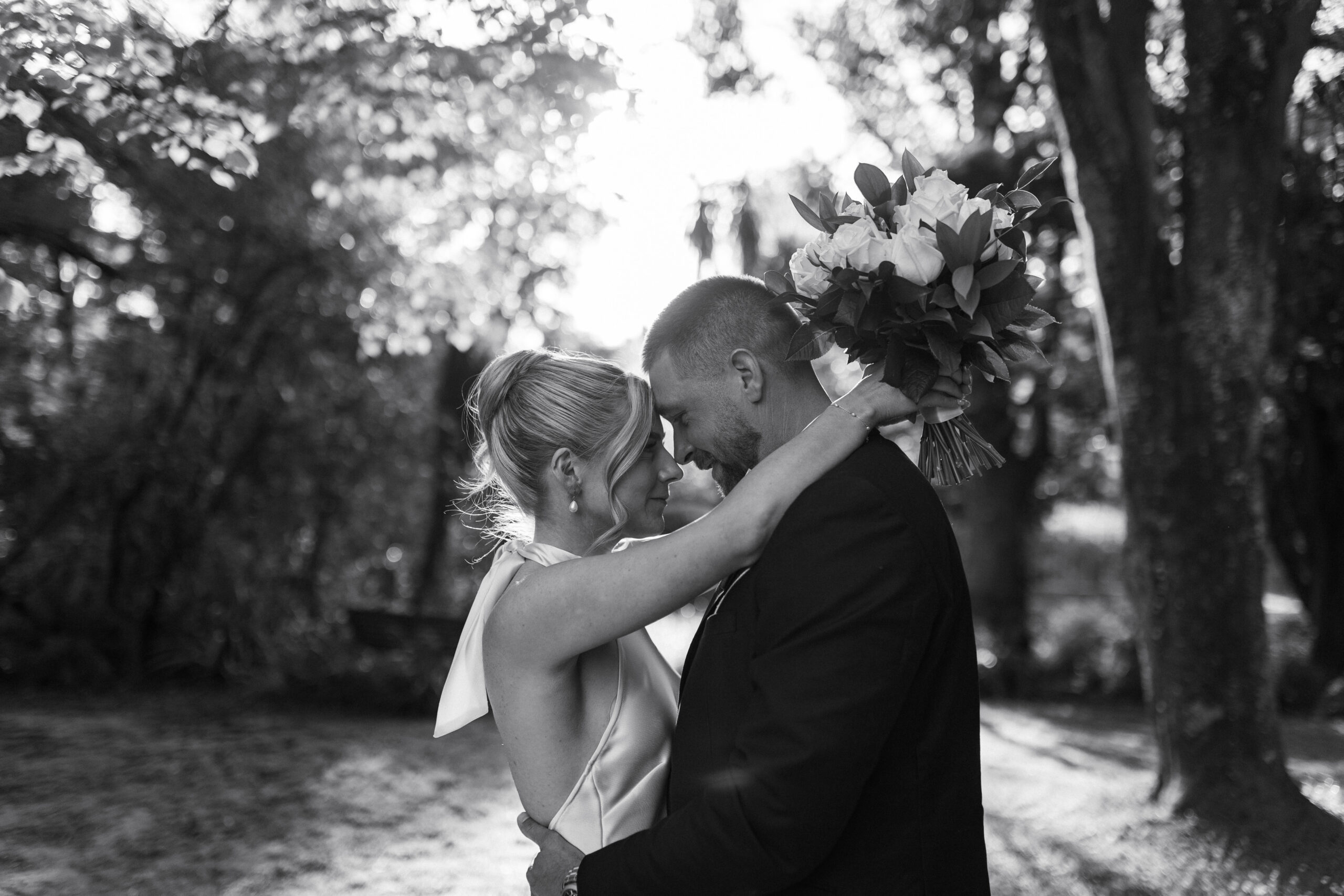 Black and white photo of a bride and groom in a tender embrace, with light filtering through trees.