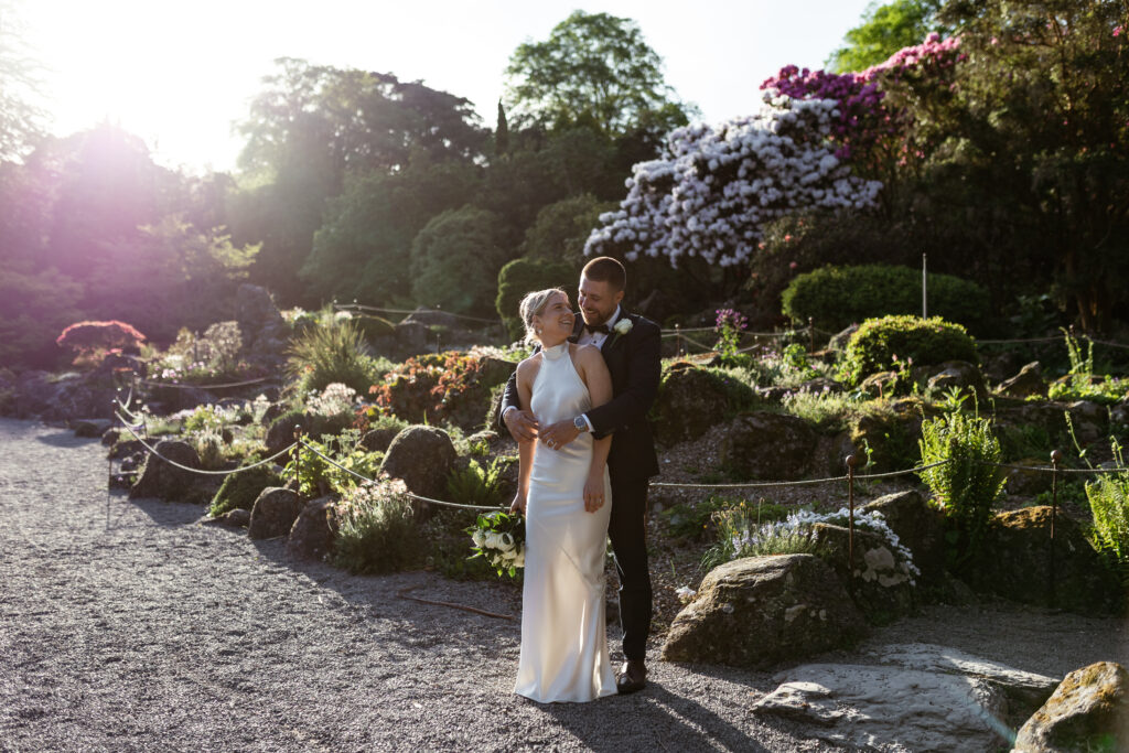 A newlywed couple embracing in one of Christchurch's signature garden settings, surrounded by lush greenery and white flowers.