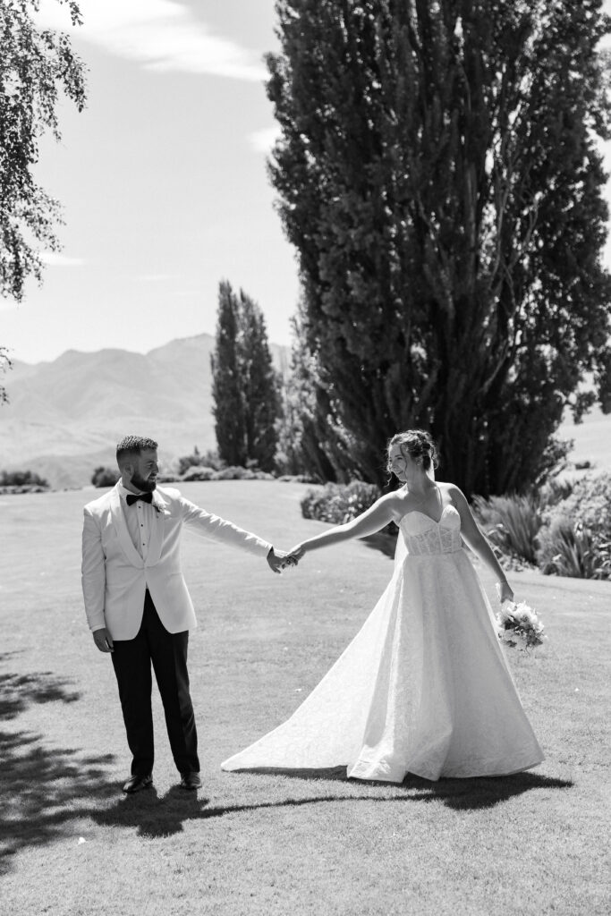 A joyful newlywed couple holding hands in a scenic Christchurch landscape, featuring tall trees and distant hills, captured in black and white.
