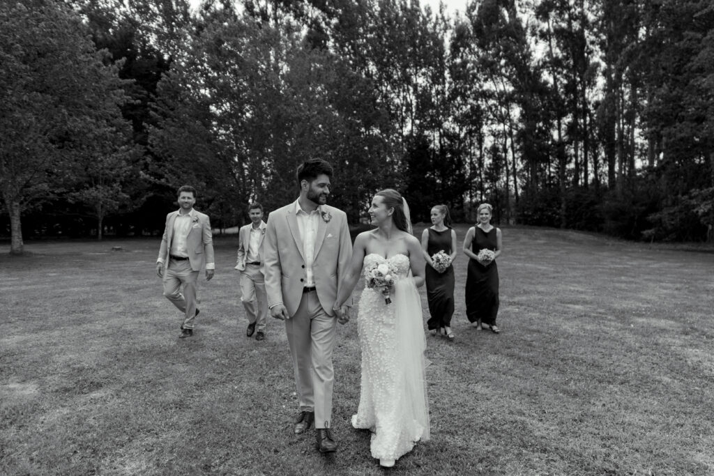 A bride and groom walking with their wedding party outdoors, captured in black and white with natural light and candid emotion.