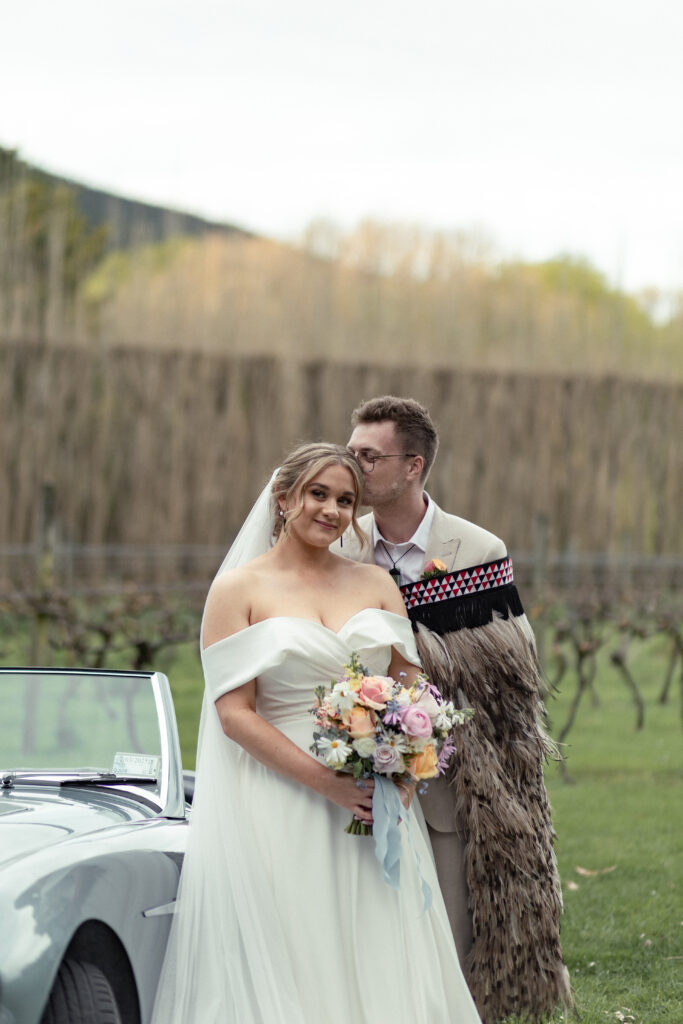 A bride and groom posing together outdoors beside a vintage car, captured in soft natural light with romantic colour tones.